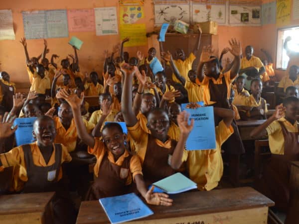 School children in a classroom sitting at desks with their hands in the air holding SPANA Humane Education work books